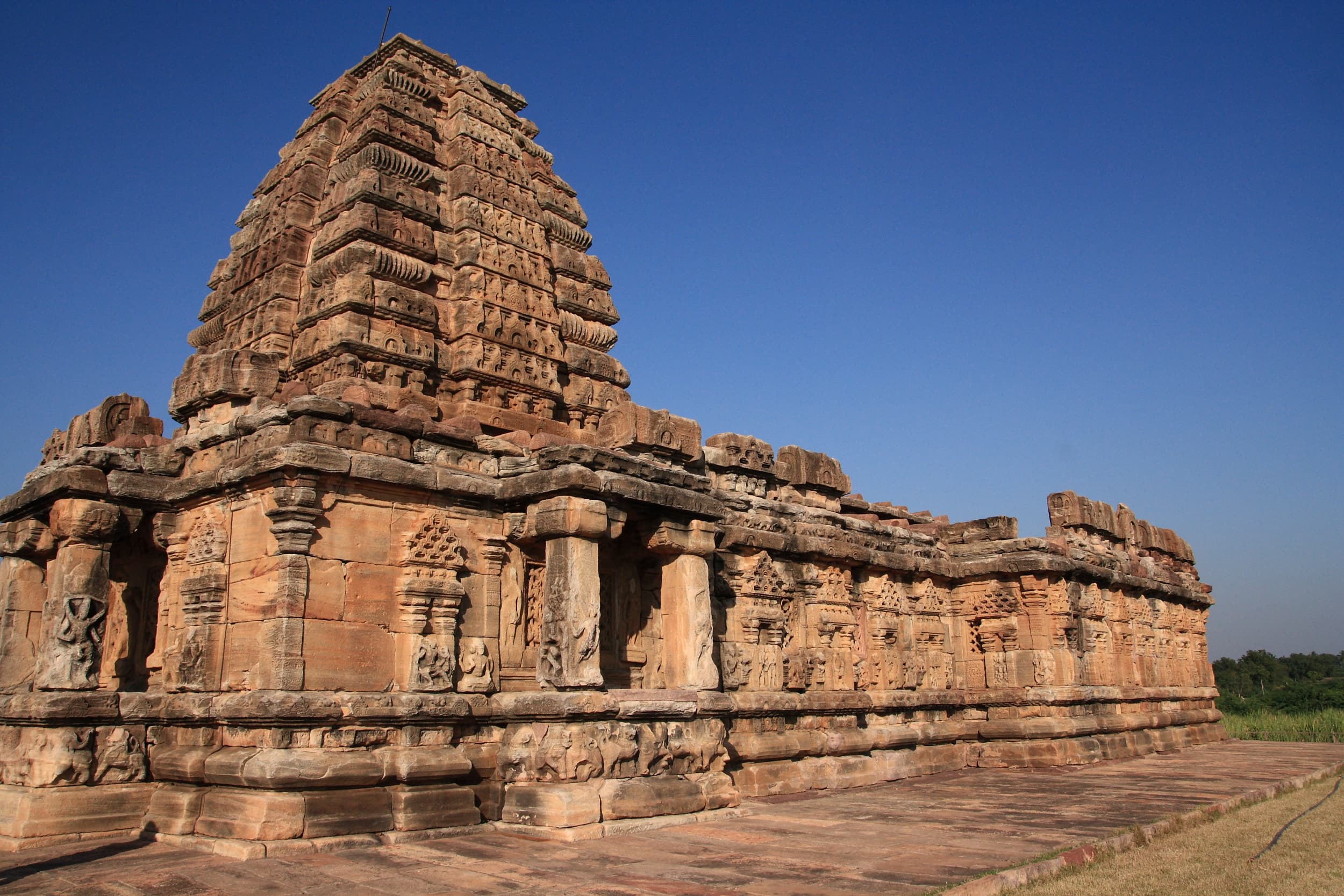 A long sandstone temple wall lined with pilasters, carved figures and small niches, with a tall tower of stacked carved tiers rising at its left end against a deep blue sky.