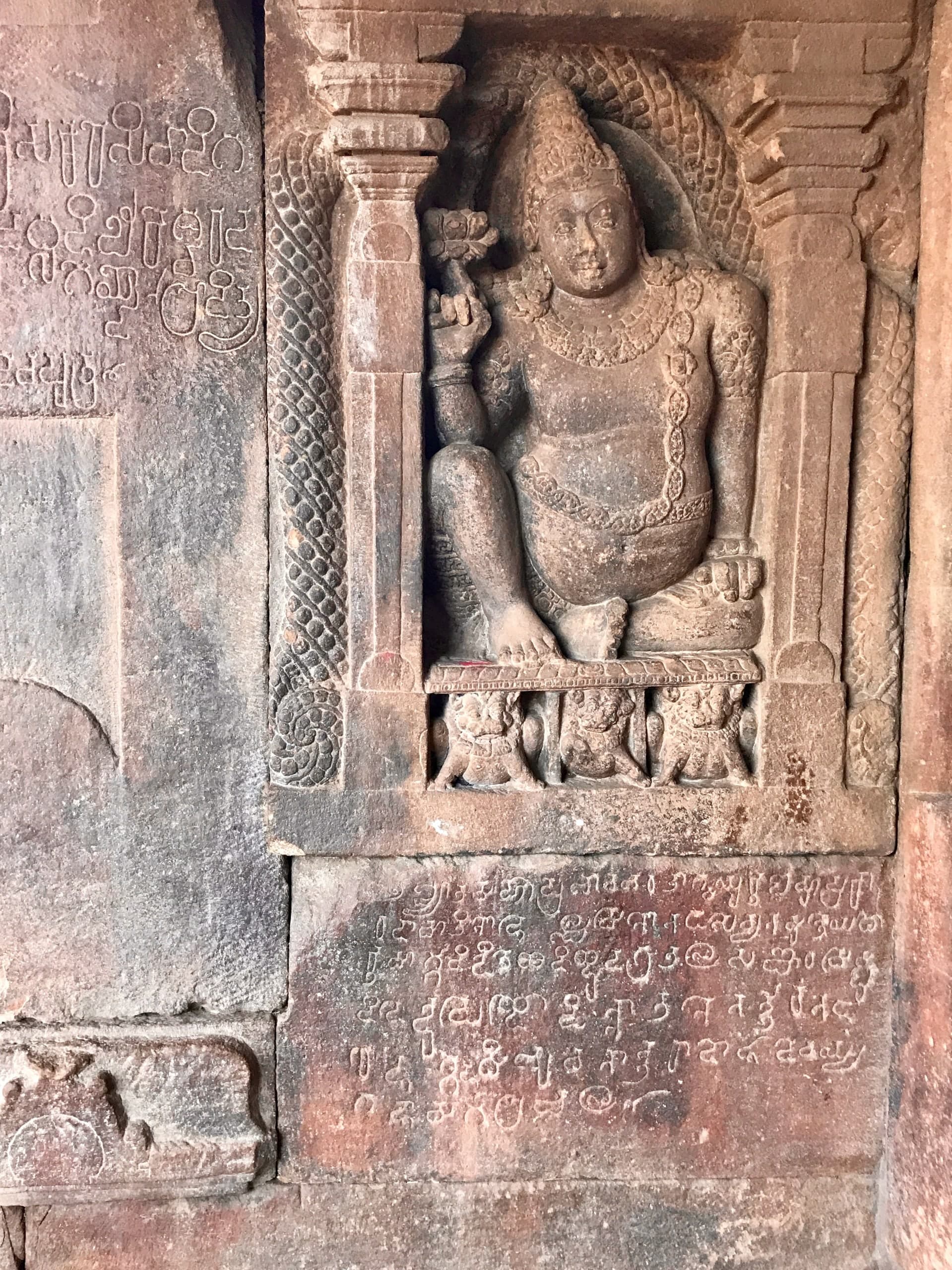 A red sandstone niche framed by pilasters holds a seated, crowned figure with one knee raised; on the panel below it and on the block to its left, lines of letters are incised into the stone.