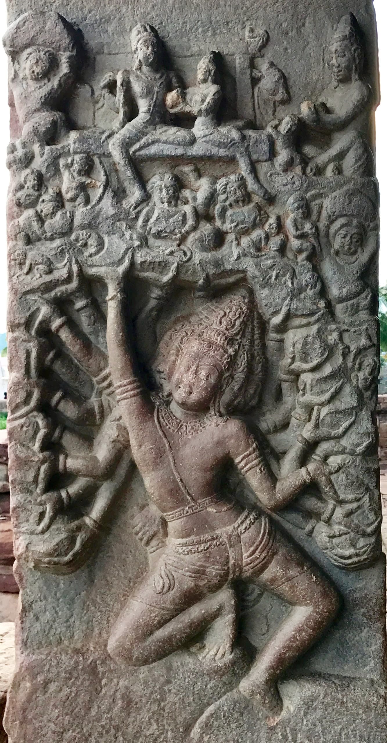A tall stone relief. At the bottom a crowned figure with many arms kneels and pushes upward with one raised arm against a crowded mass of small figures, above which a seated couple and attendants sit on a ledge.