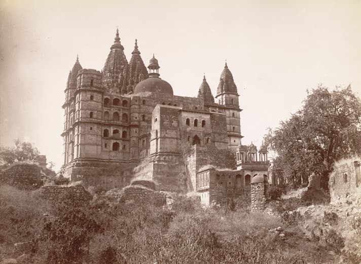 A sepia photograph of a massive temple on a high stone base, crowned by tall clustered spires, corner towers and a central dome, seen from below across rough ground with low ruined walls, scrub and a large tree at right.