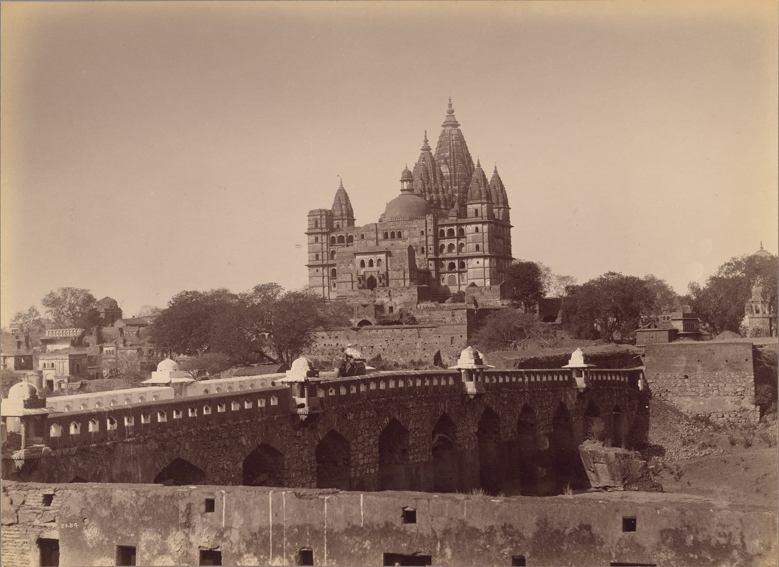 A sepia photograph of a tall temple crowned by a cluster of spires and a dome, standing on high ground above trees and low houses. In front, a long arched stone bridge with a pierced parapet and small domed kiosks is crossed by a rider on an elephant.