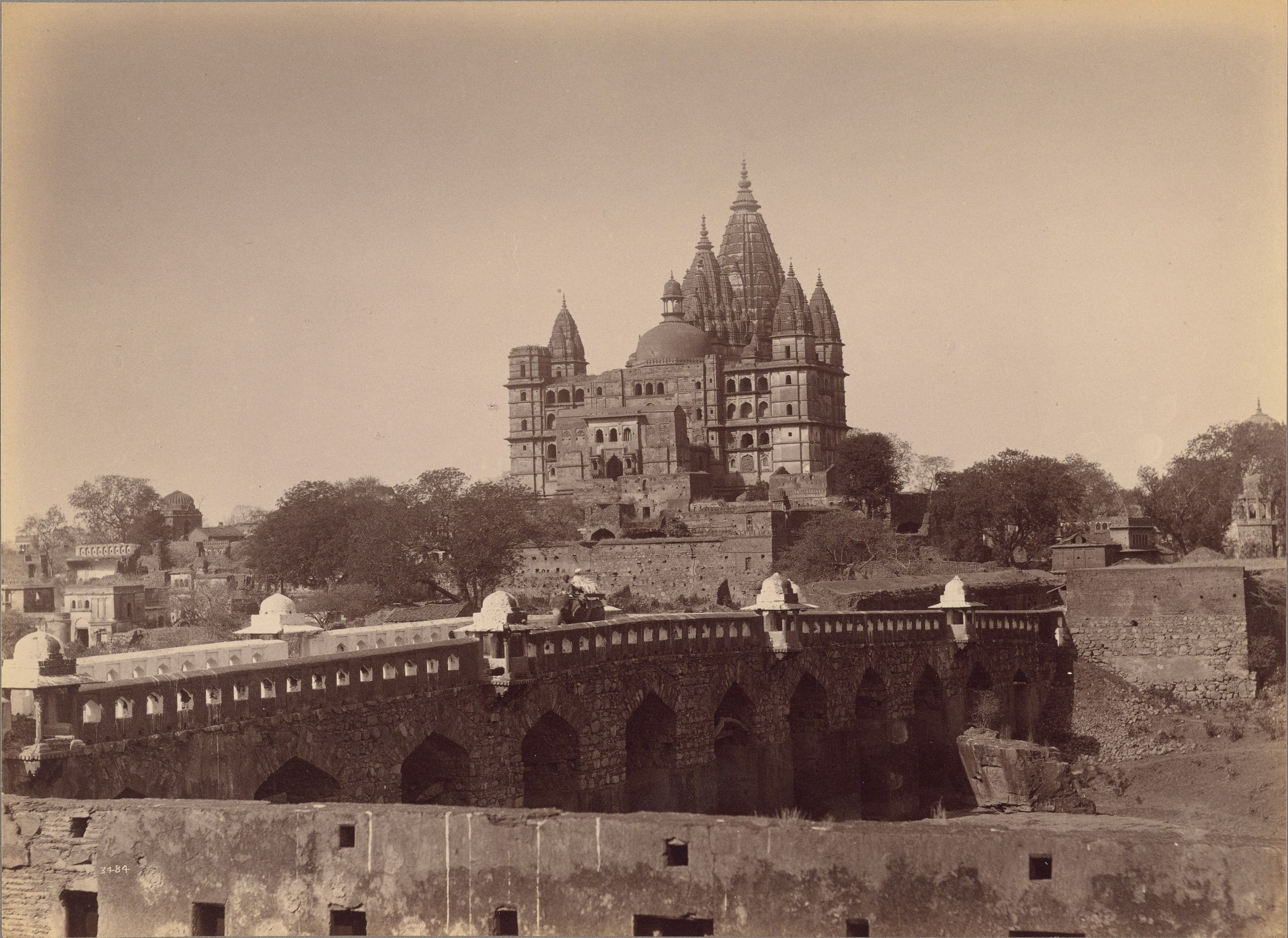 A sepia photograph of a tall temple crowned by a cluster of spires and a dome, standing on high ground above trees and low houses. In front, a long arched stone bridge with a pierced parapet and small domed kiosks is crossed by a rider on an elephant.