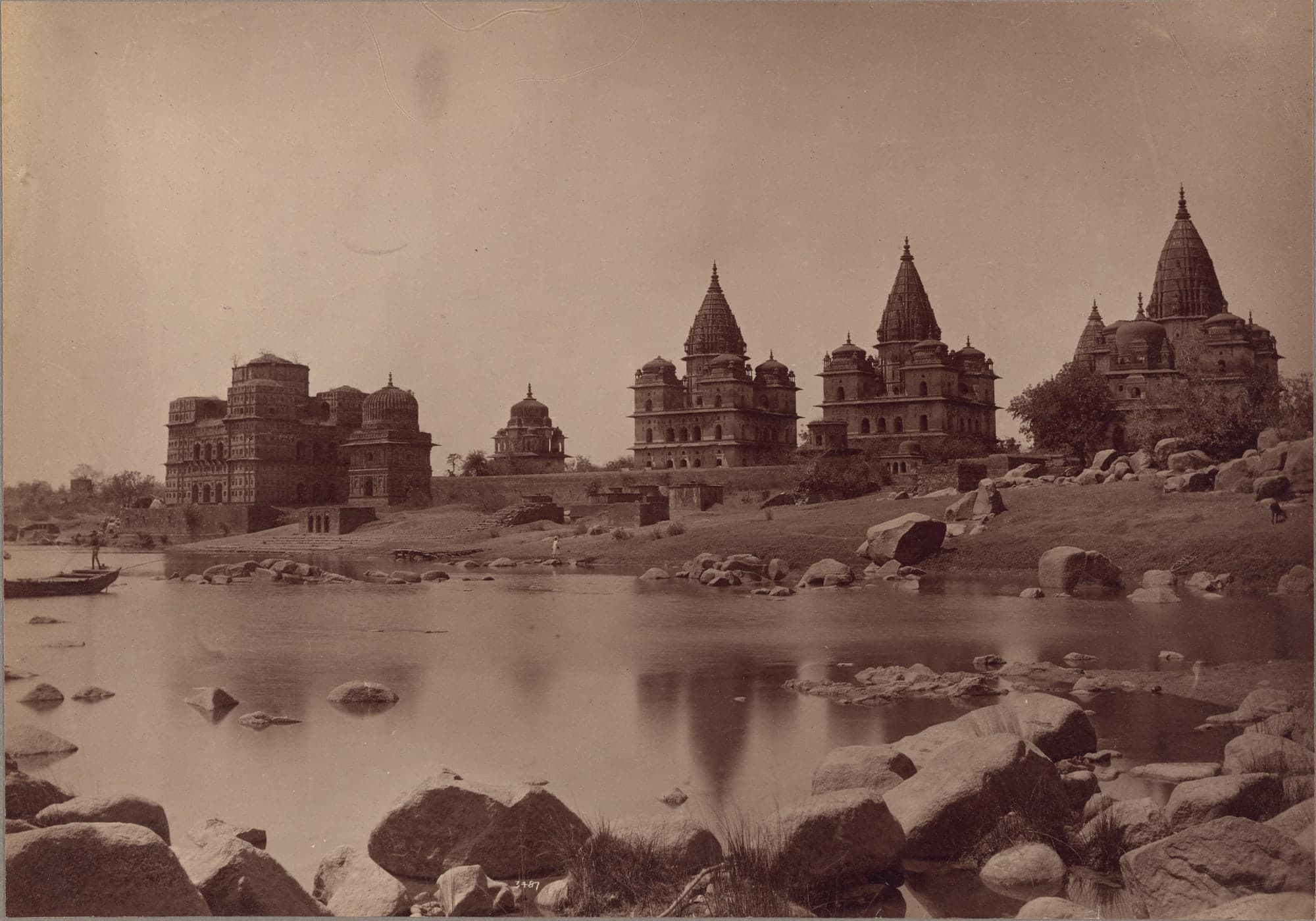 A sepia photograph of a row of stone cenotaphs on a riverbank, several crowned by tall spires and domes, one at left a squarer block with plants on its roof. A man poles a boat at left; boulders and still water fill the foreground.