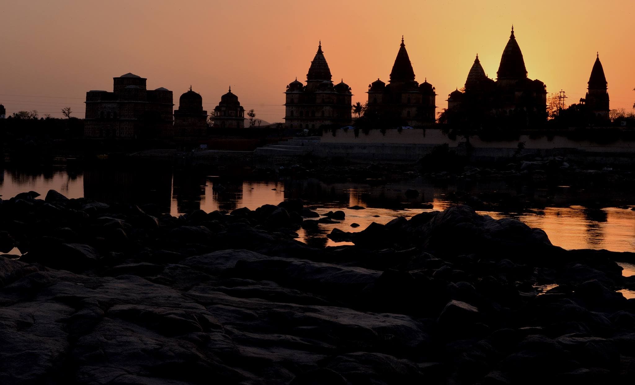 The silhouettes of domed and spired cenotaphs against an orange sunset sky, reflected in the river, with dark boulders in the foreground.