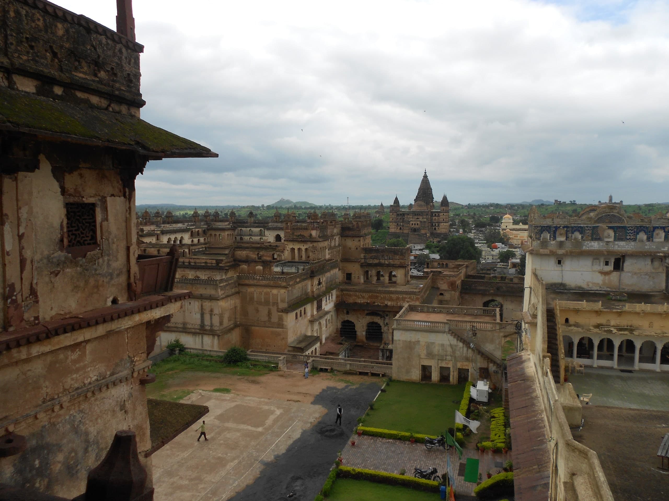 A high view from a weathered palace wall over courtyards, arcades and rows of small domed kiosks. Beyond them a tall temple with a central spire and corner towers rises over the town and green countryside under a cloudy sky.