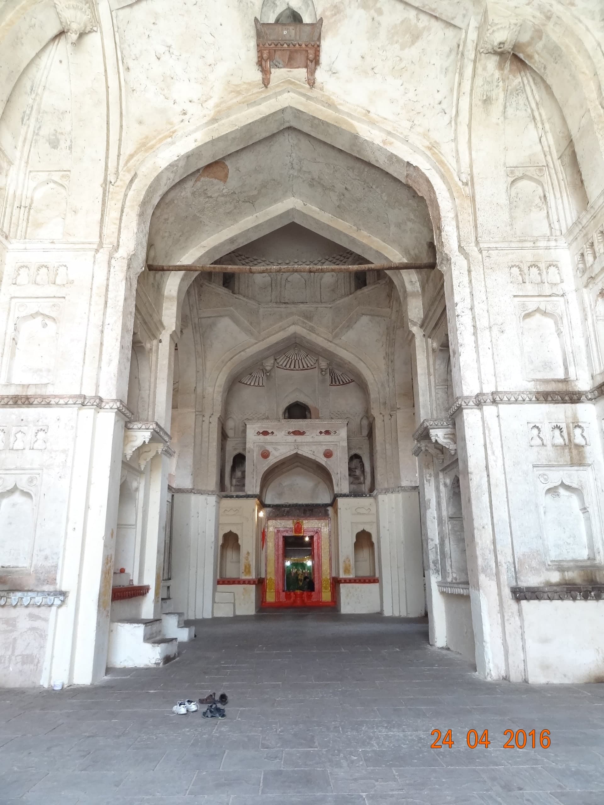 A tall whitewashed interior with nested pointed arches and plain walls set with small niches. At the far end of the stone-floored hall, a doorway framed in red and gold is lit from within. A few pairs of shoes lie on the floor in the foreground.