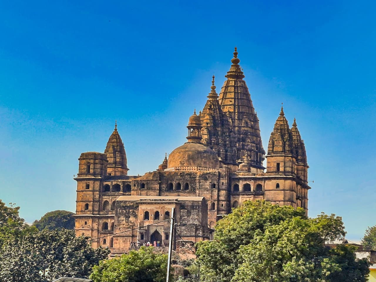 A tall weathered stone temple with a large central spire, smaller spires at its corners and a dome, rising above green trees against a deep blue sky.