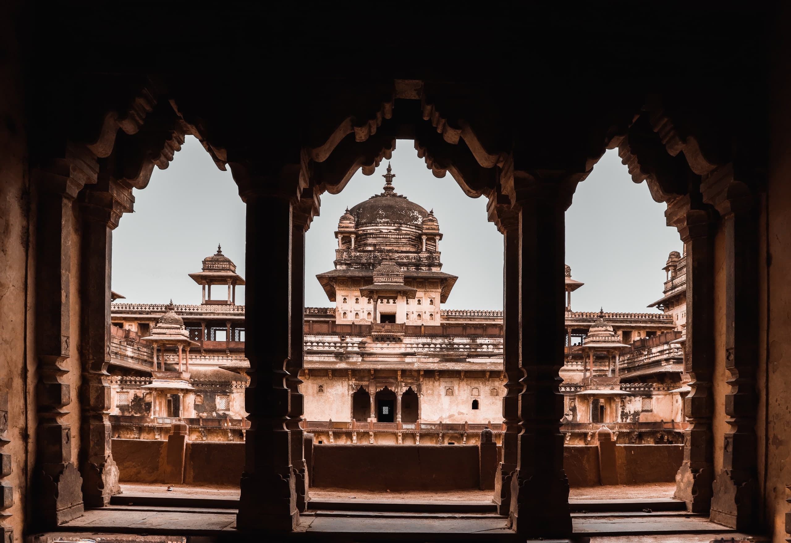 Seen through three cusped stone arches, a palace courtyard with tiered balconies, small domed kiosks on slender pillars, and a large central dome.