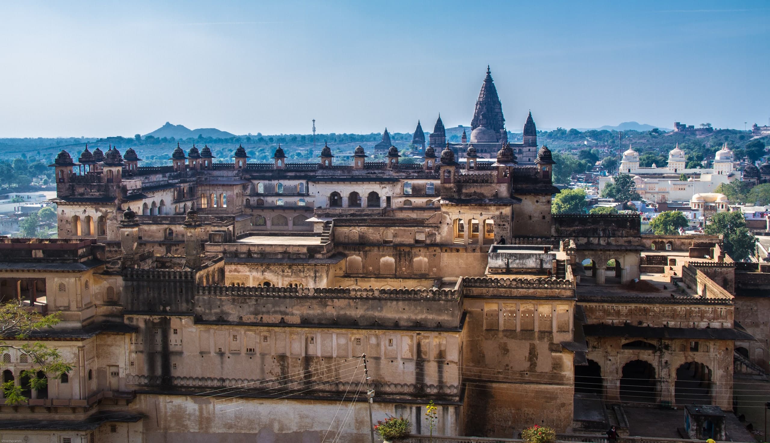 A weathered stone palace of courtyards and arcades topped by rows of small domed kiosks fills the foreground. Behind it rise the dark spires and dome of a tall temple, and to the right stands a cream-coloured building with white domes among the town’s houses.