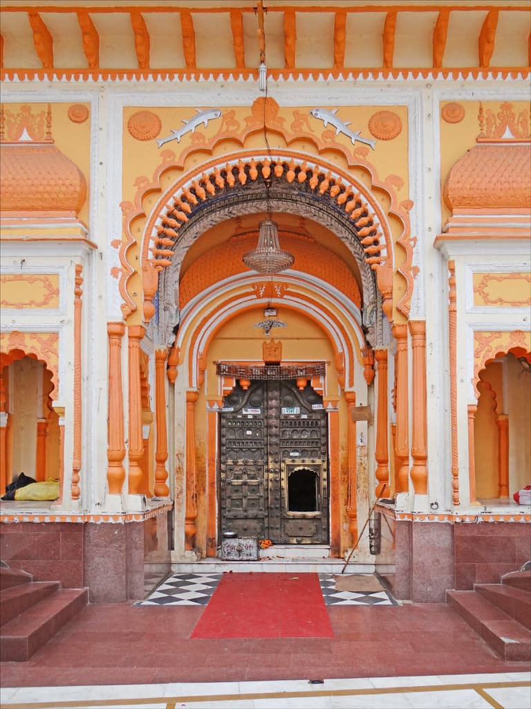 A tall cusped archway painted orange, cream and white, with two fish motifs above it and a chandelier hanging inside, frames a closed pair of studded metal doors. A red carpet runs to the doors over a chequered floor.