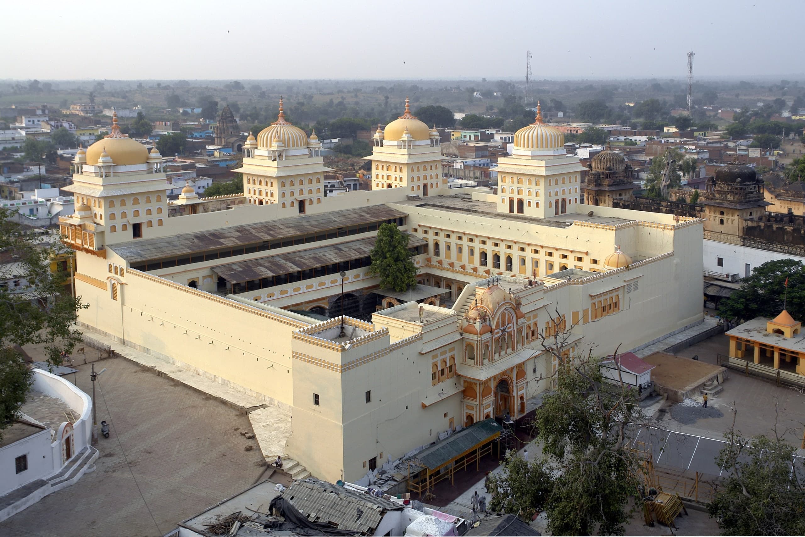 A high view of a large cream-coloured building with four tall ribbed domes on square towers, set around a courtyard partly roofed with sheets. An ornate orange-and-cream gateway stands at the front, facing an open paved square.