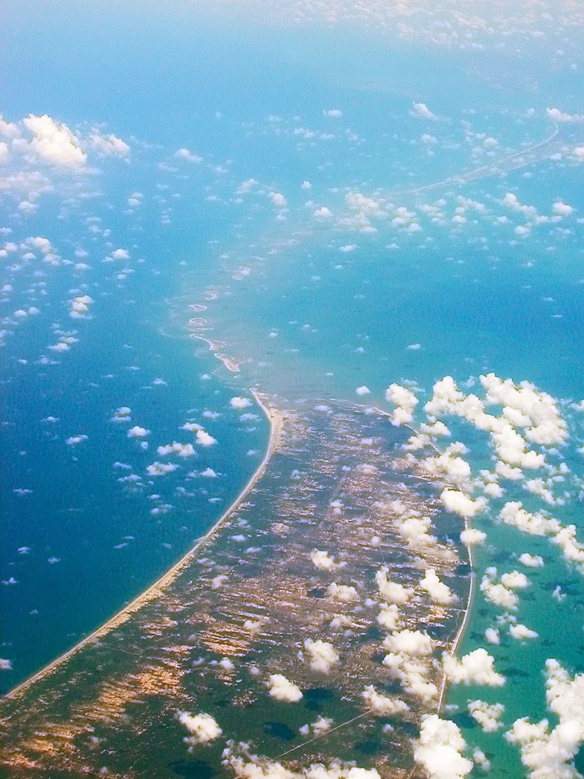 Aerial photograph of a narrow, curving line of pale sandbanks and shallow water stretching across a turquoise sea from a large landmass in the lower half of the frame, with scattered white clouds below the aircraft.