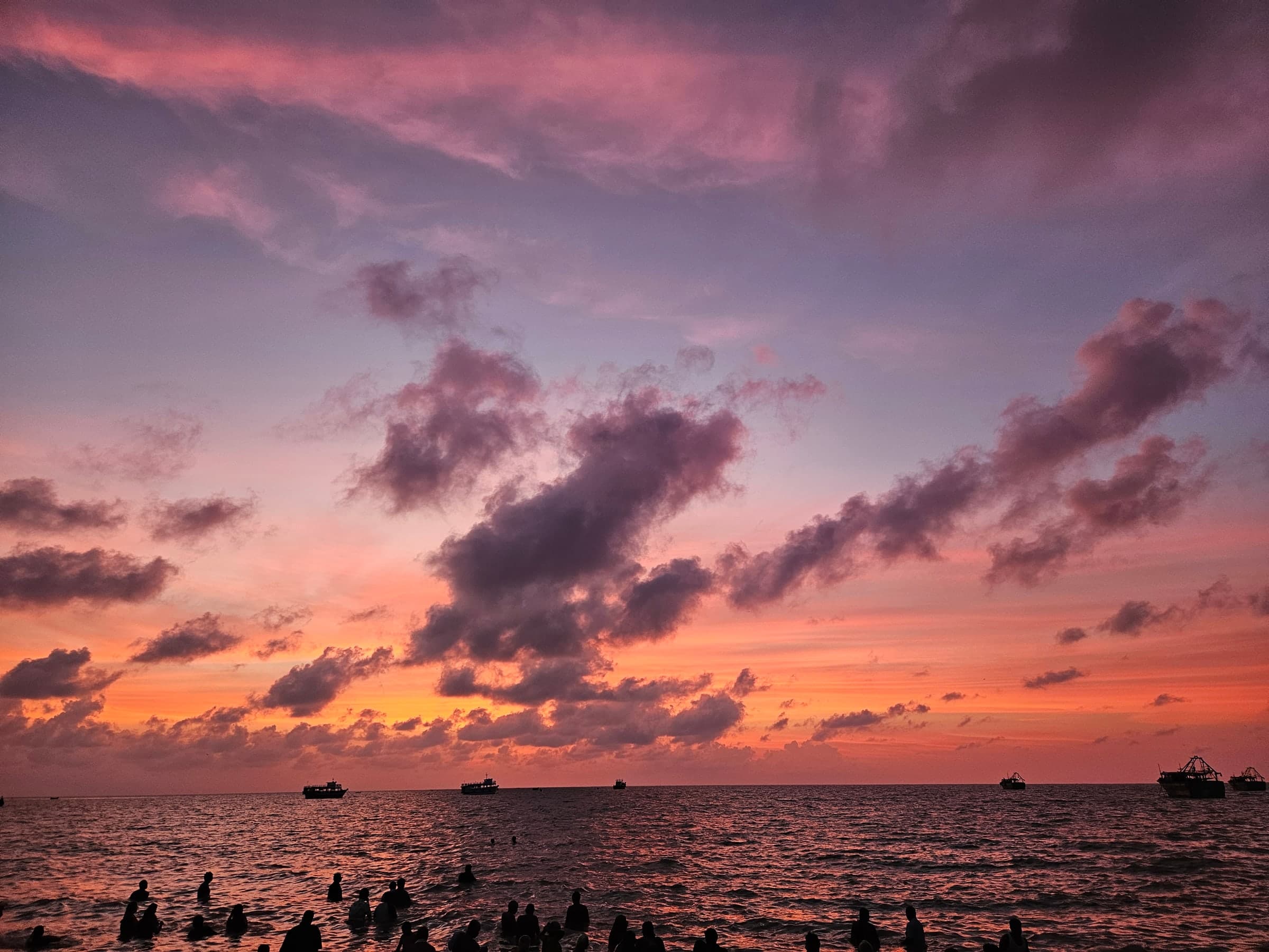 A vivid pink and purple dawn sky over a calm sea. Small boats sit on the horizon, and the dark silhouettes of people standing waist-deep in the water line the foreground.