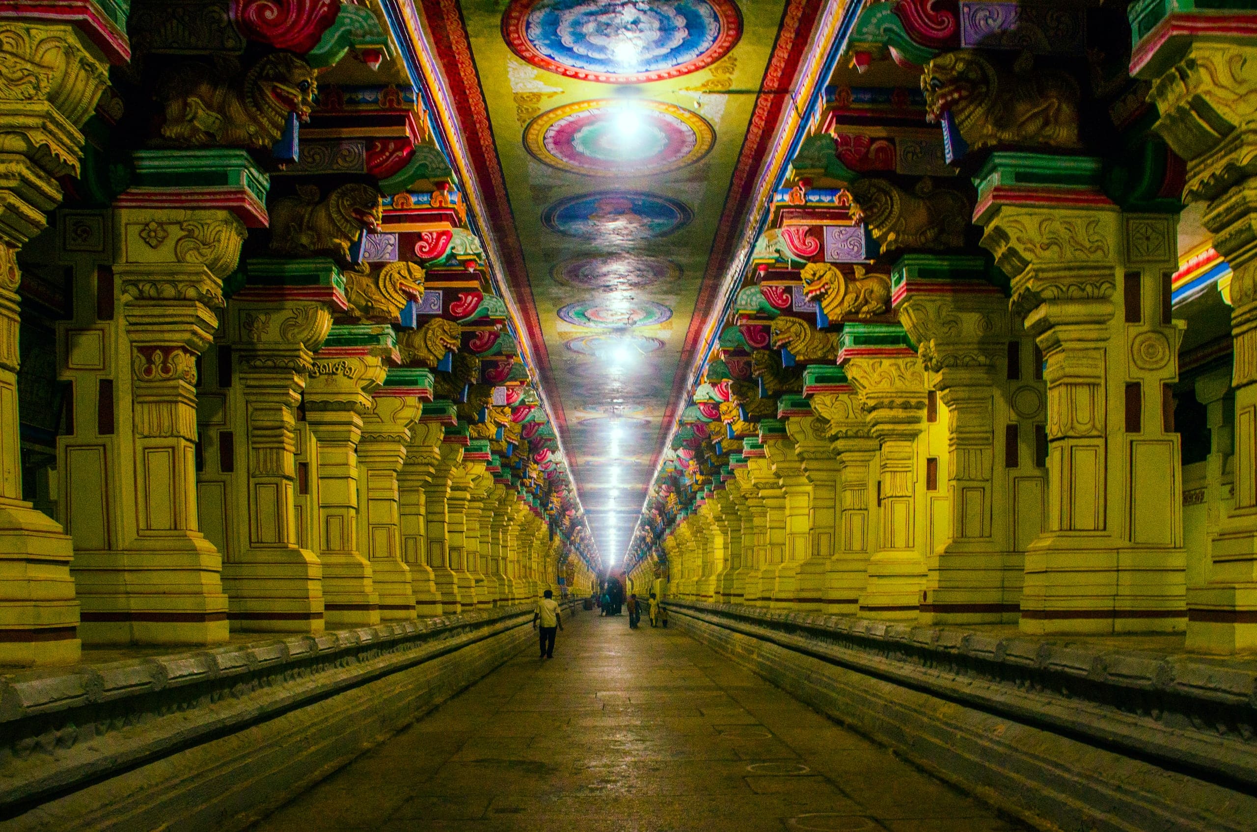 A long, straight corridor running into the distance between rows of massive carved pillars painted yellow, red and green, under a ceiling of painted circular medallions. A few people walk far down the passage.