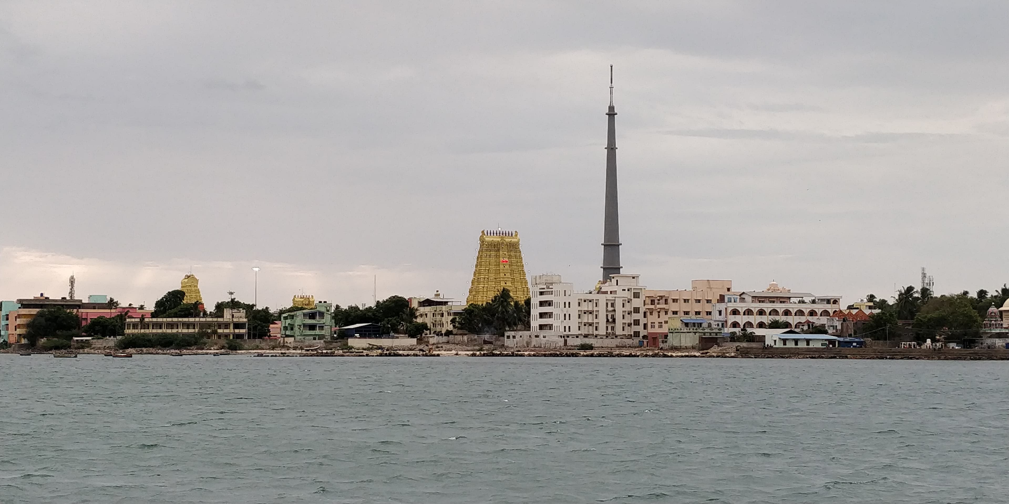 A wide view across grey sea water to a low town on the shore. Temple towers, the tallest golden-yellow, rise above the buildings, beside a slender communications mast, under an overcast sky.