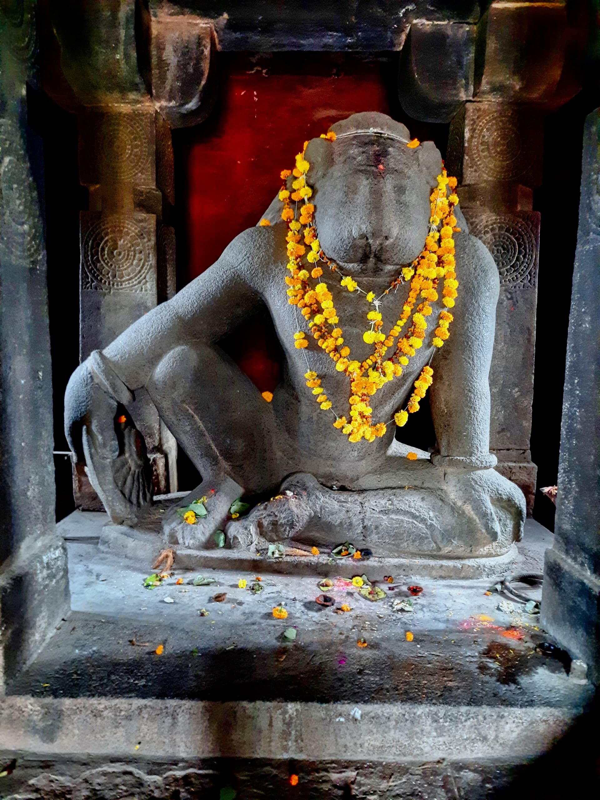 A large seated stone figure with a lion’s head, one knee drawn up with an arm resting across it, hung with orange and yellow marigold garlands, in a dim shrine between carved stone pillars. Flower offerings lie on the pedestal.