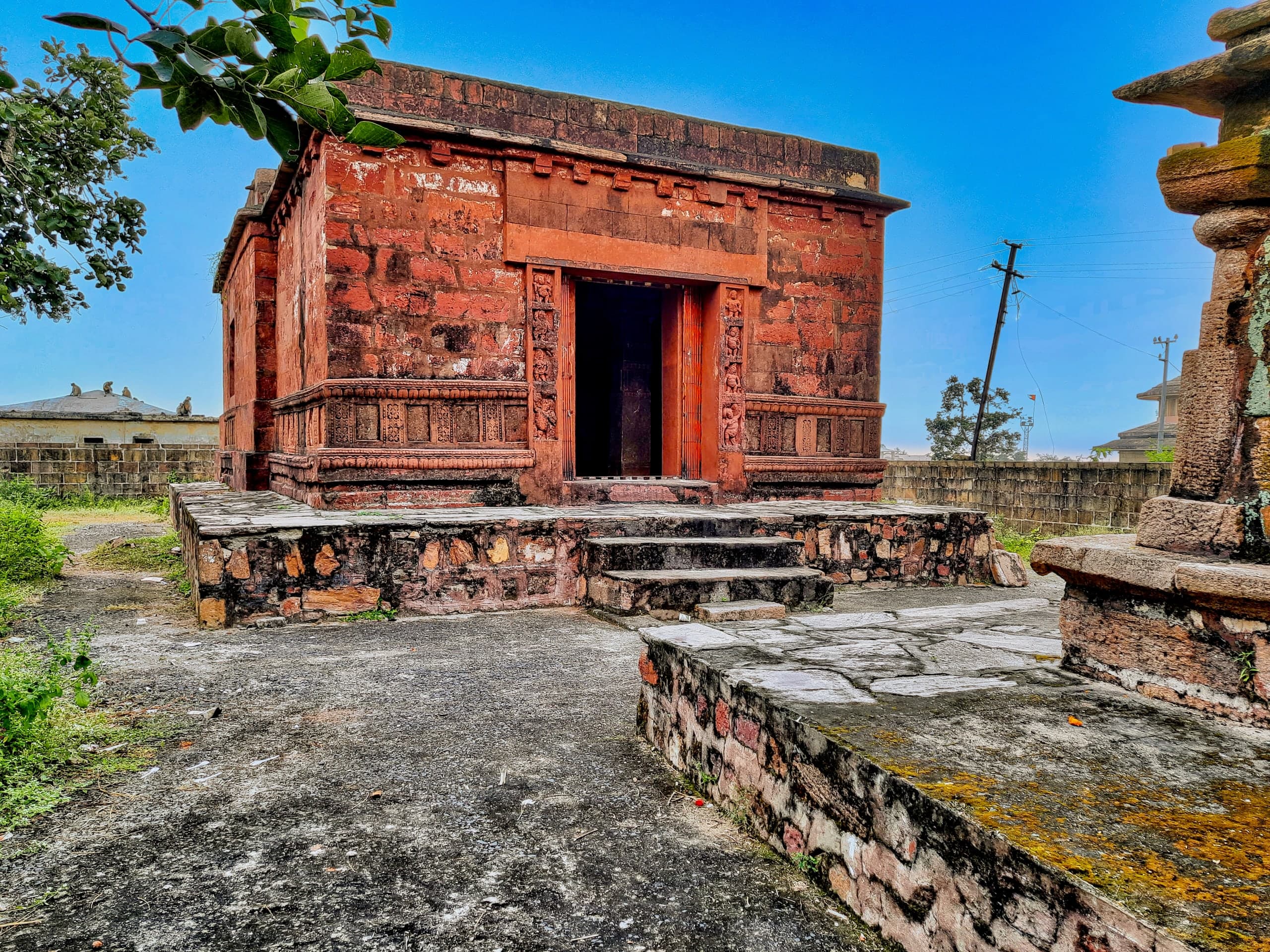 A small, flat-roofed temple of weathered red sandstone on a low rubble-stone platform with steps, its dark doorway framed by carved jambs and a carved band along the plinth. Trees, a wall and utility poles stand around it.