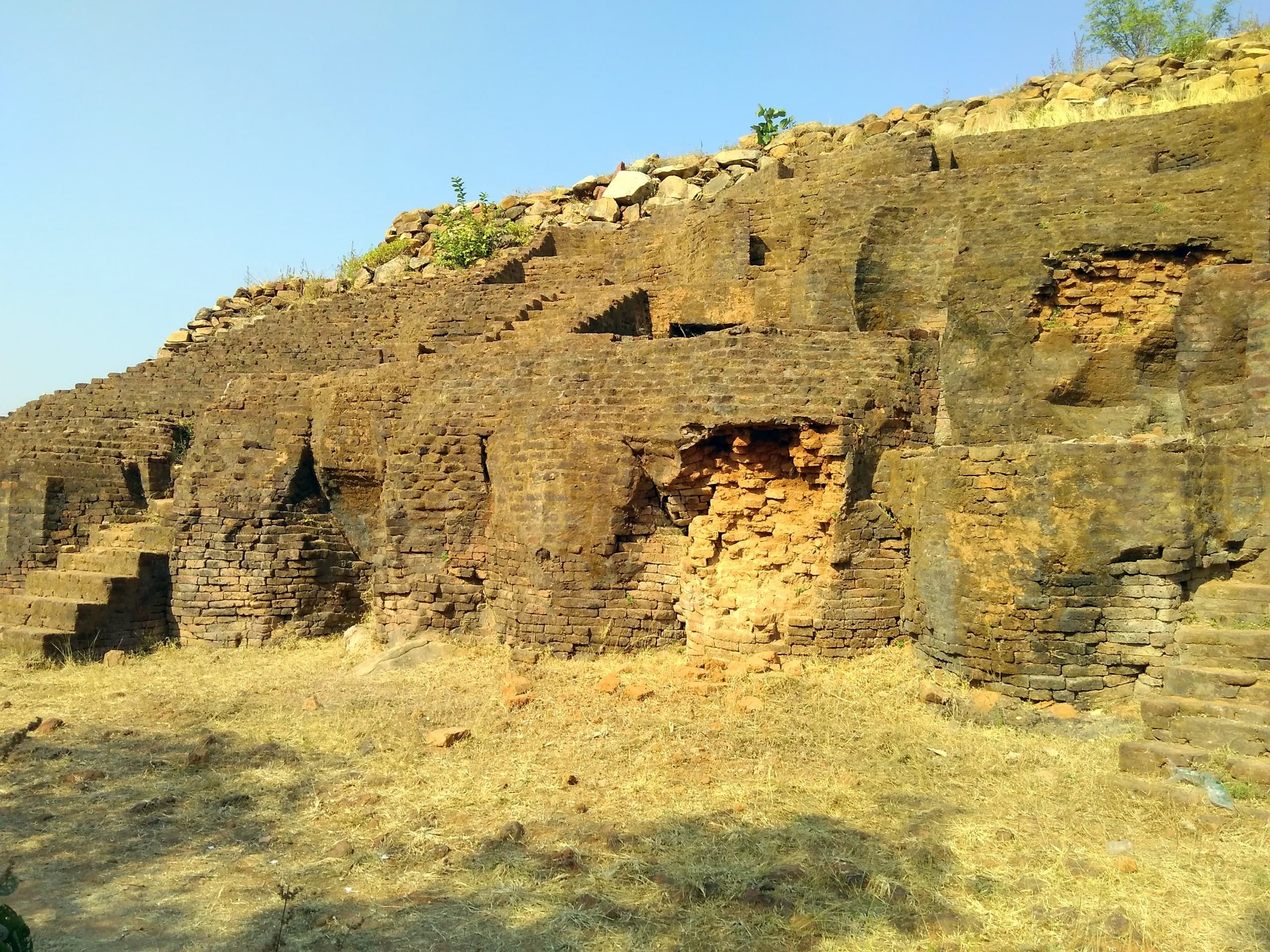 Excavated walls of dark brick rising in steps and terraces up the side of a mound, with patches of exposed orange brick, dry grass in the foreground and clear sky above.