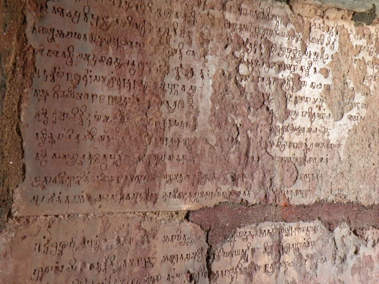 Close view of a reddish stone surface in several slabs, carved with rows of rounded, looped letters. Parts of the surface are flaked away and the slabs are cracked and broken at the edges.