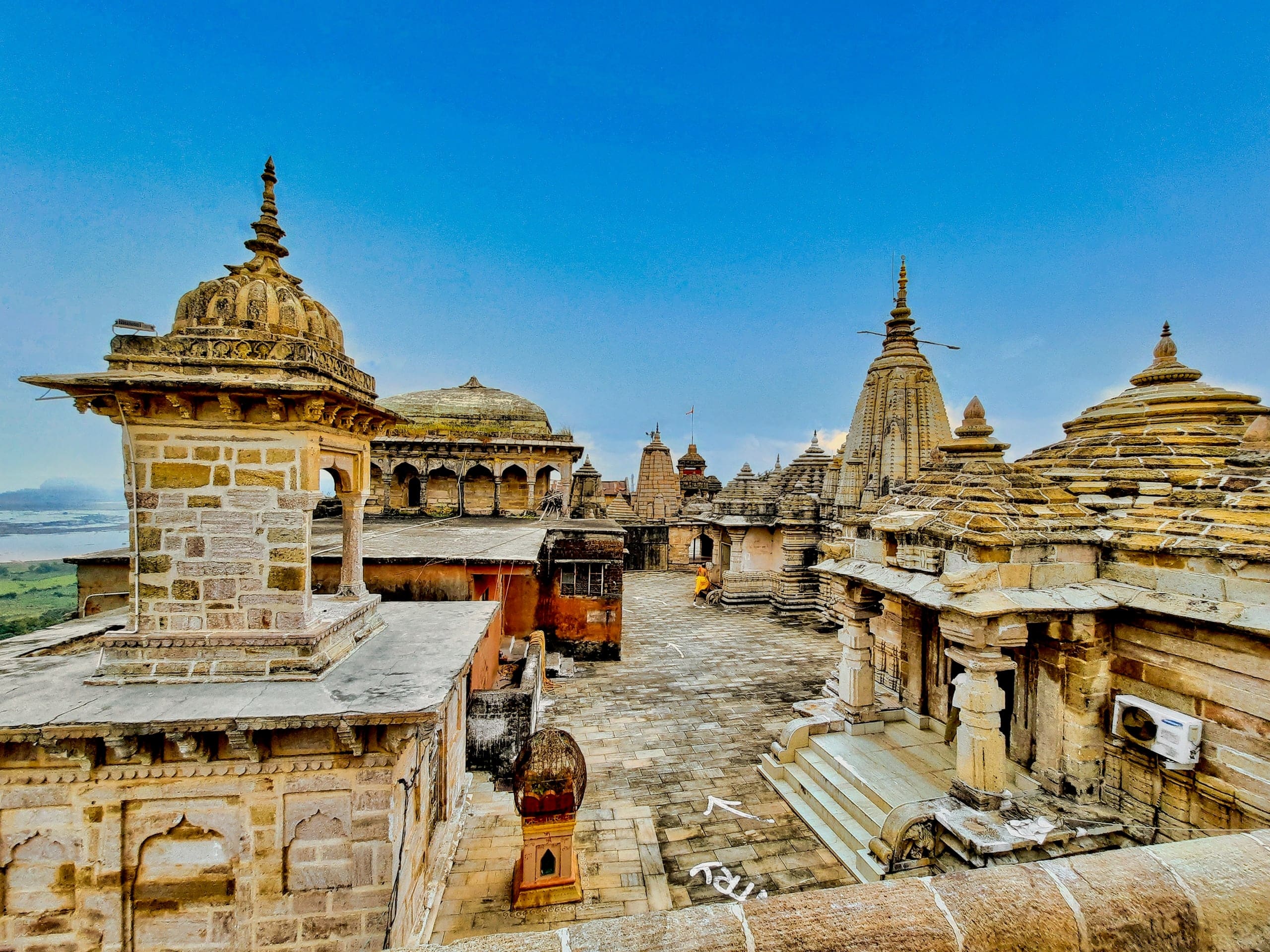 A wide-angle view across stone rooftops to a paved court ringed by sandstone shrines with tall carved spires, a domed pavilion and a smaller domed kiosk at left, under a deep blue sky. A plain and a stretch of water lie far below at left.