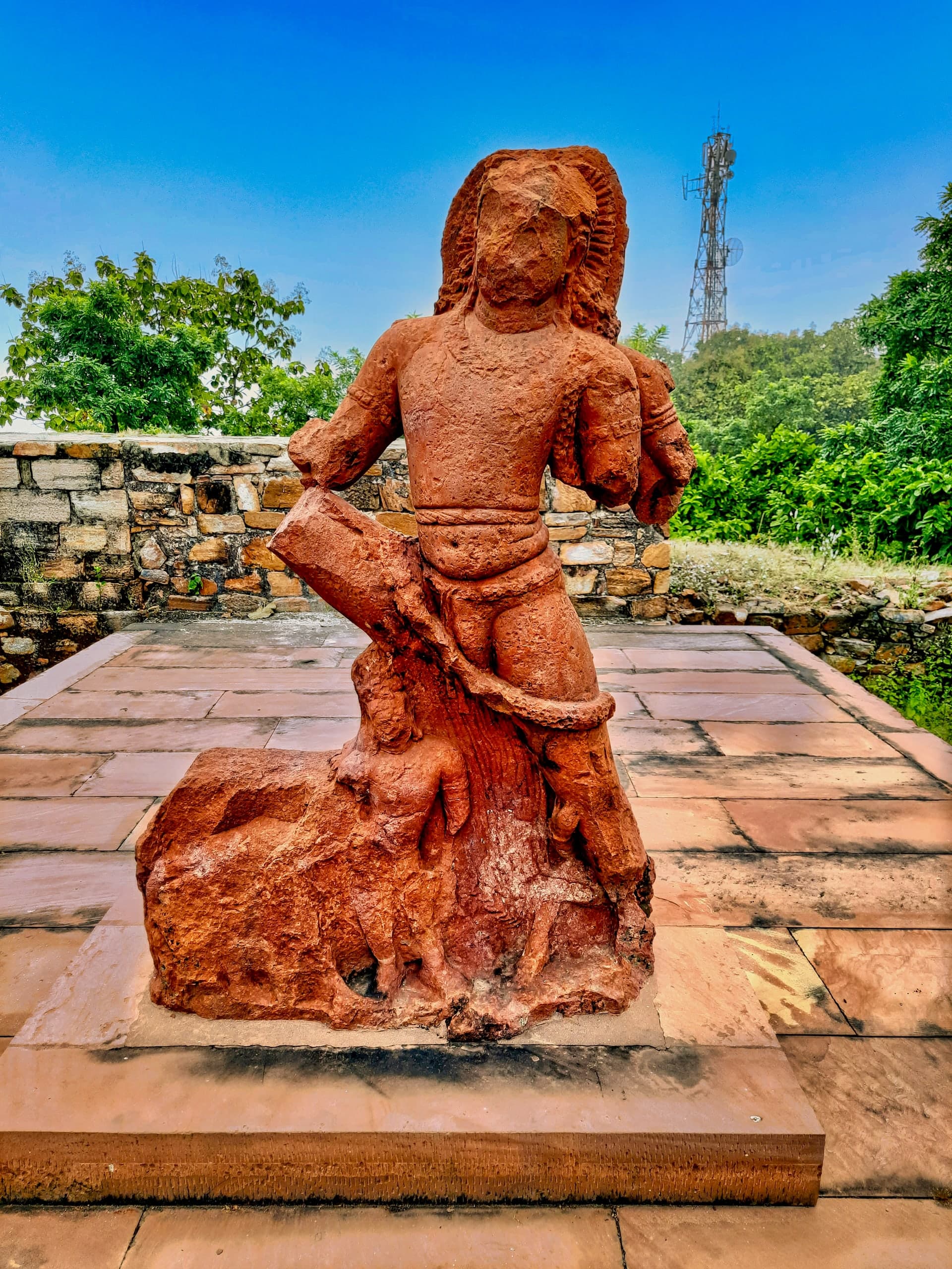 A battered standing figure carved in red sandstone on a modern stone base, its face worn away and its arms broken, with a smaller carved figure near its feet. A telecommunications mast rises behind the trees.