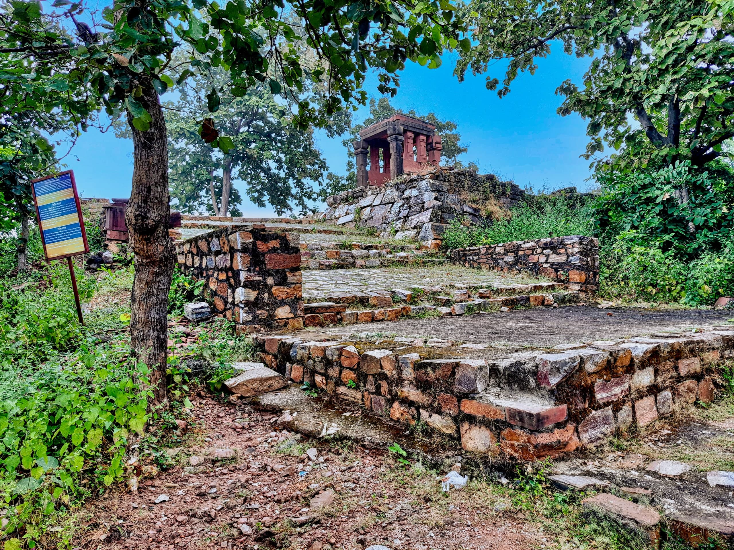 Rubble-stone terraces and low walls climb among trees to a small pillared pavilion of red sandstone at the top. An information board on a post stands at the left.