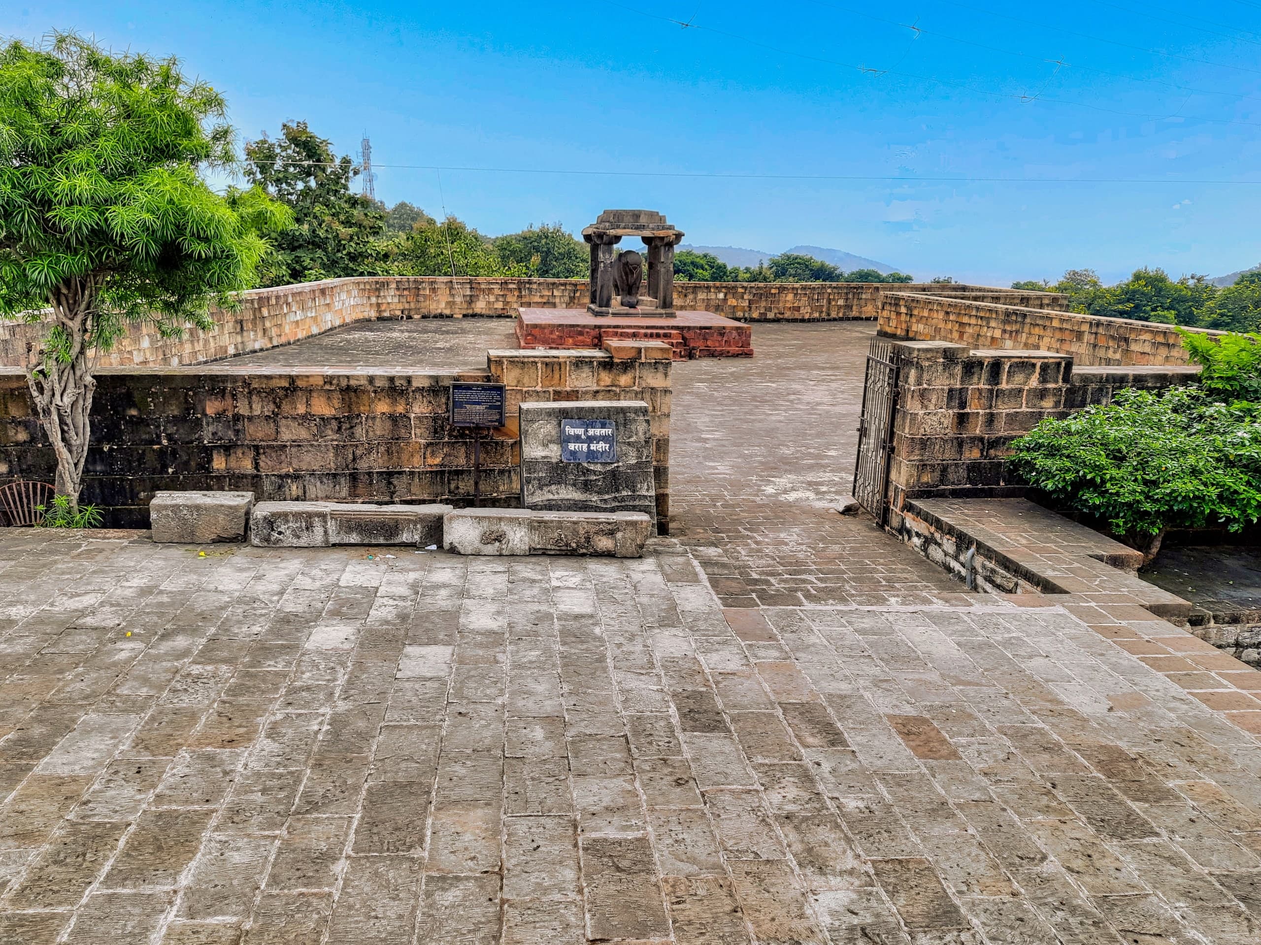 A broad stone-paved terrace with low sandstone walls leads to a small open pavilion of four pillars on a red platform, with a dark stone animal figure inside. A blue sign in Devanagari stands by the wall, with trees and hills beyond.